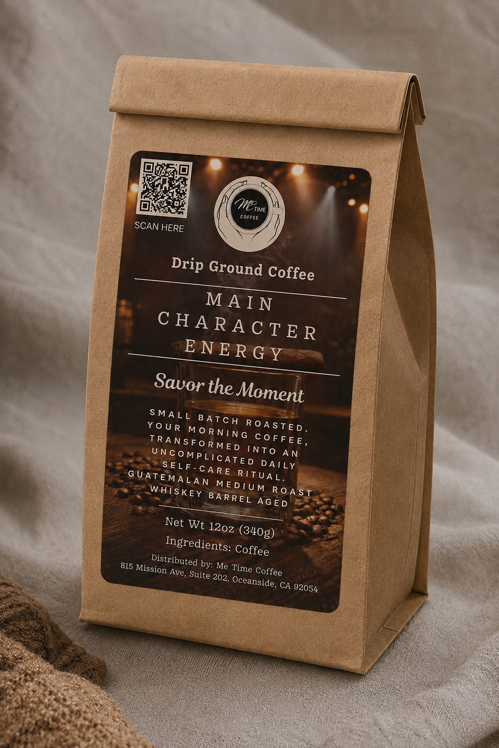 A kraft paper coffee bag standing upright on a draped fabric surface. The front label reads 'Drip Ground Coffee' and 'Main Character Energy' with a coffee-themed dark label and a QR code near the top left.