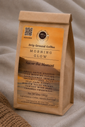 A kraft paper coffee bag labeled 'MORNING GLOW' sits on a soft beige fabric surface. The bag is made of brown kraft paper with a large orange and brown label showing coffee flavor and roast details, plus a QR code and small logo.